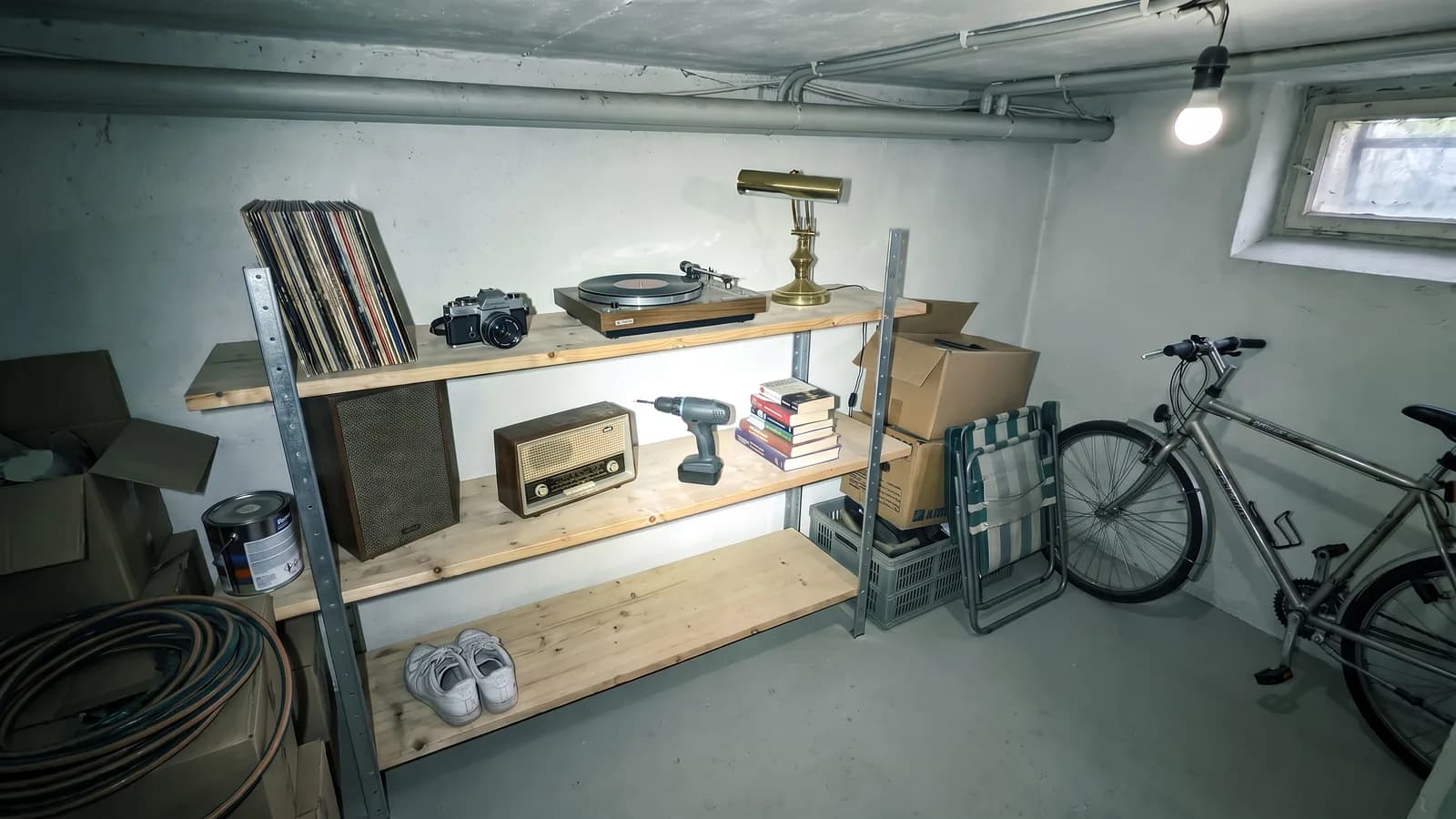 Ein Kellerraum mit Rohren an der Decke und einer nackten Glühbirne: auf einem einfachen Regal stehen eine Spiegelreflexkamera, ein Plattenspieler, eine Messinglampe, ein altes Radio, Sneaker, ein Akkuschrauber und Bücher; daneben Kartons, ein Gartenschlauch und ein Campingstuhl, an der Wand lehnt ein Fahrrad.